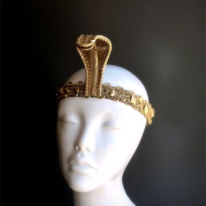 May include: A gold-colored cobra headdress with intricate detailing. The cobra rises from a decorative headband with circular embellishments. The headdress is displayed on a white mannequin head against a dark background.