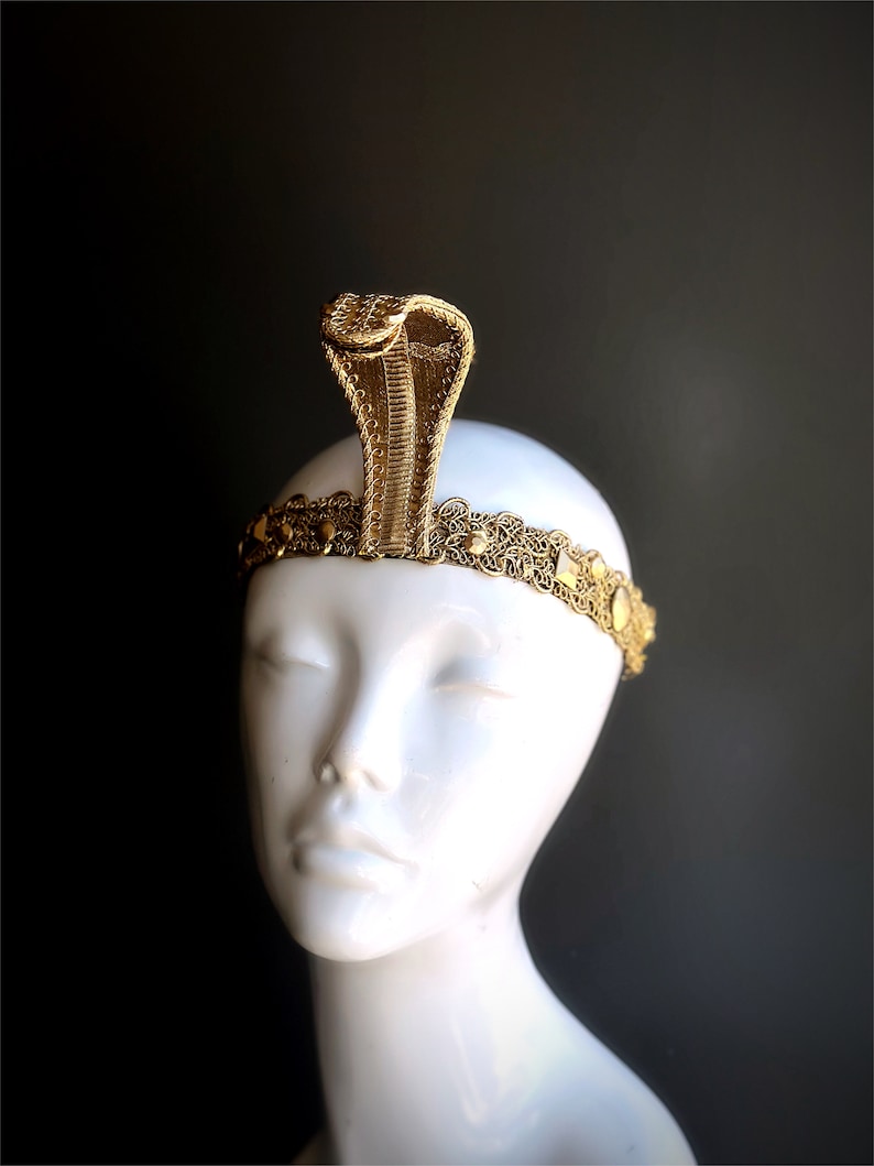 May include: A gold-colored cobra headdress with intricate detailing. The cobra rises from a decorative headband with circular embellishments. The headdress is displayed on a white mannequin head against a dark background.