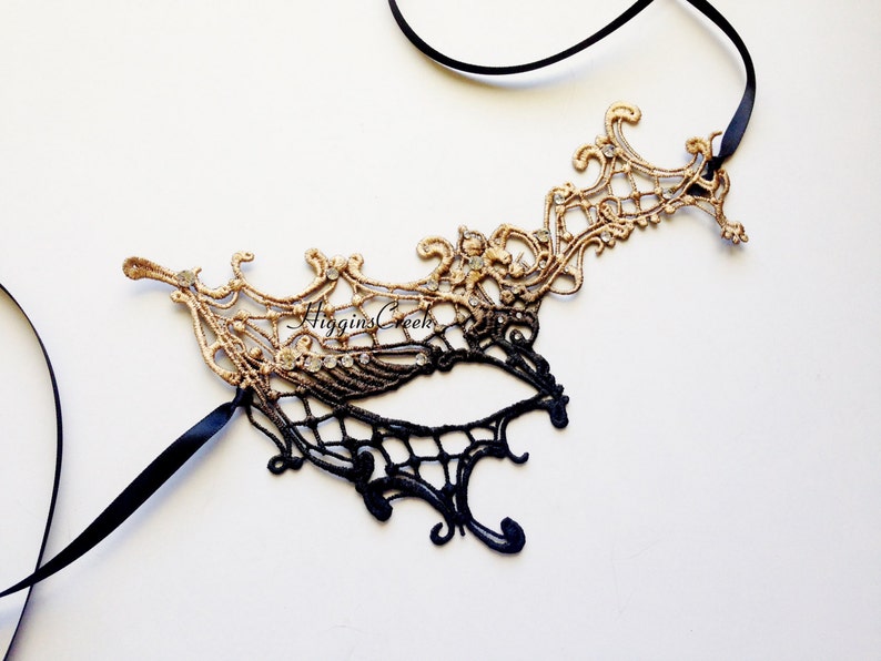 May include: A black and gold masquerade mask with intricate lace detailing. The mask is attached to a black ribbon.