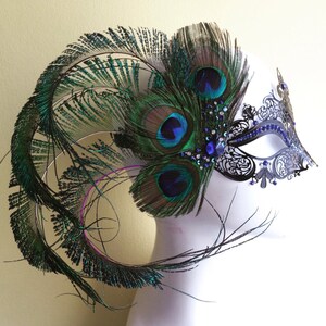 May include: A black masquerade mask with intricate lace detailing and blue rhinestones. The mask is adorned with a large peacock feather plume in shades of green and blue.