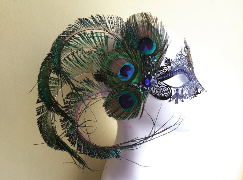 May include: A black masquerade mask with intricate lace detailing and blue rhinestones. The mask is adorned with a large peacock feather plume in shades of green and blue.