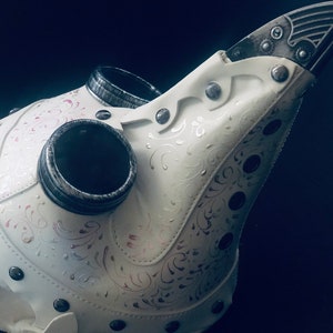 May include: A white plague doctor mask with silver accents and decorative floral patterns. The mask features a long, beak-like nose, two eye holes, and two circular air vents. Rivets secure the leather pieces together.