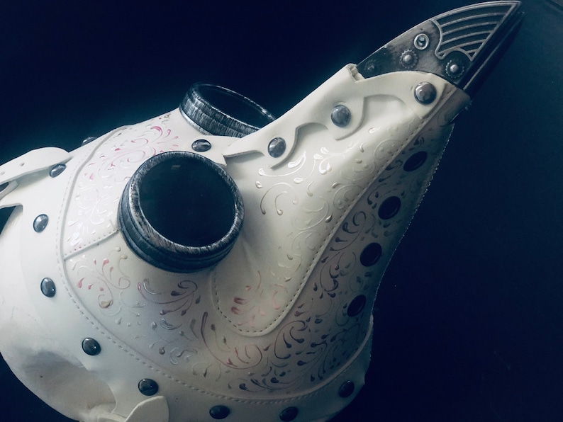May include: A white plague doctor mask with silver accents and decorative floral patterns. The mask features a long, beak-like nose, two eye holes, and two circular air vents. Rivets secure the leather pieces together.