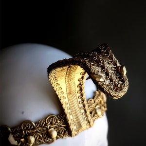 May include: A gold-colored cobra headpiece with intricate detailing. The headpiece features a cobra head with a raised hood and a decorative band with pearls. The headpiece is likely for costume or theatrical use.