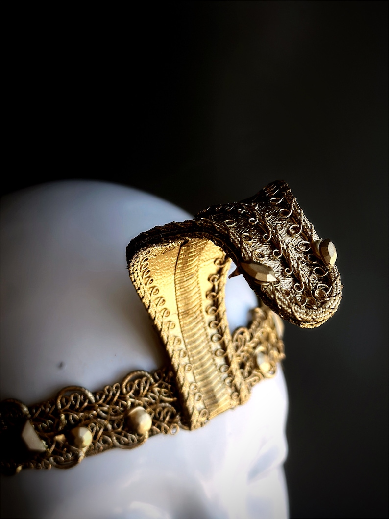 May include: A gold-colored cobra headpiece with intricate detailing. The headpiece features a cobra head with a raised hood and a decorative band with pearls. The headpiece is likely for costume or theatrical use.
