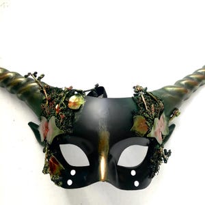 May include: A black and gold masquerade mask with curved, textured horns. The mask is decorated with green and red foliage, and has eye holes and two sets of three white dots. A black strap is attached to the mask.