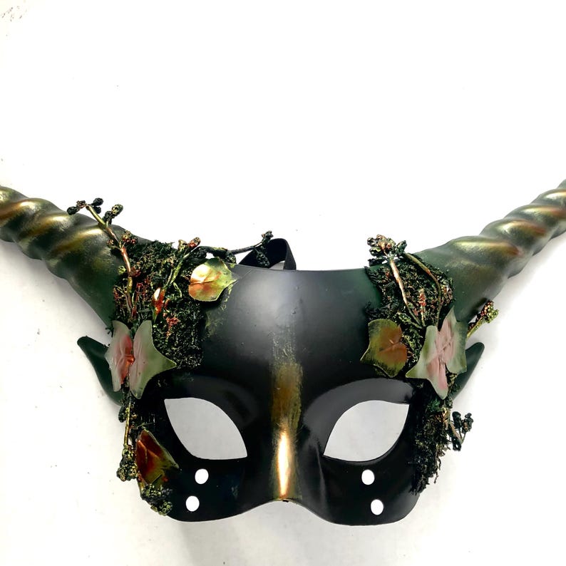 May include: A black and gold masquerade mask with curved, textured horns. The mask is decorated with green and red foliage, and has eye holes and two sets of three white dots. A black strap is attached to the mask.