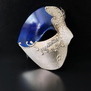 Könnte beinhalten: Eine venezianische Maske in Blau und Silber. Der obere Teil ist glänzend blau mit einem feinen Muster, während die untere Hälfte eine strukturierte, rissige Silberoberfläche aufweist. Noten sind auf der Maske abgebildet.