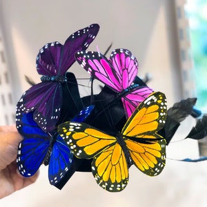 May include: A black headband with five colorful butterfly embellishments. The butterflies are purple, pink, yellow, and blue.