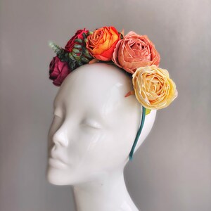 May include: A floral headband with orange, pink and yellow fabric flowers. The headband is teal green and is worn on a white mannequin head.
