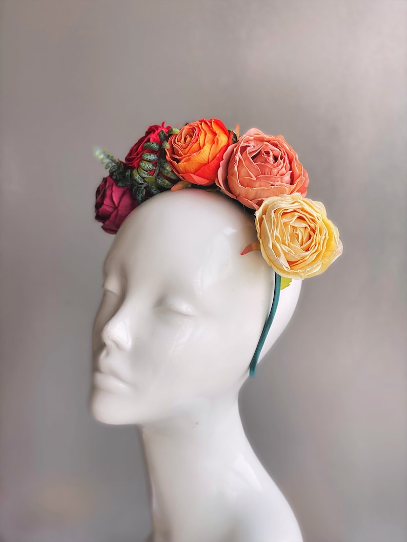 May include: A floral headband with orange, pink and yellow fabric flowers. The headband is teal green and is worn on a white mannequin head.