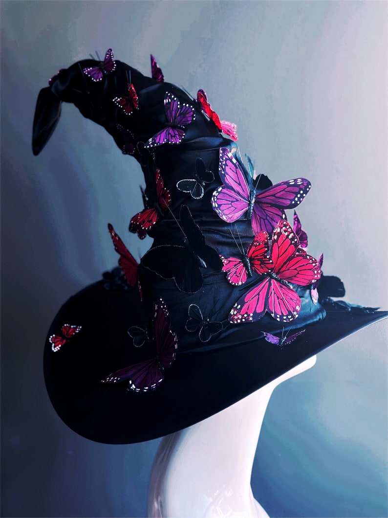 May include: A black witch hat with a tall, pointed crown adorned with pink and red butterflies. The hat is made of a soft, velvety fabric and has a wide brim.