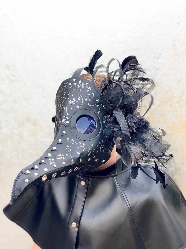 May include: A black leather plague doctor mask with a silver floral design and a black feather trim. The mask has a long beak and a round eye piece.