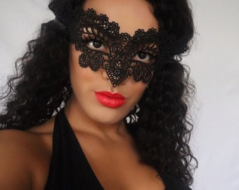 May include: A woman with long, curly black hair wearing a black lace masquerade mask. The mask covers her eyes and has a delicate, intricate design.