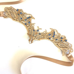 May include: A gold lace headpiece adorned with blue gemstones. The headpiece features an intricate, openwork pattern and is attached to a beige ribbon. This item is likely intended for a special event, such as a wedding or formal occasion.