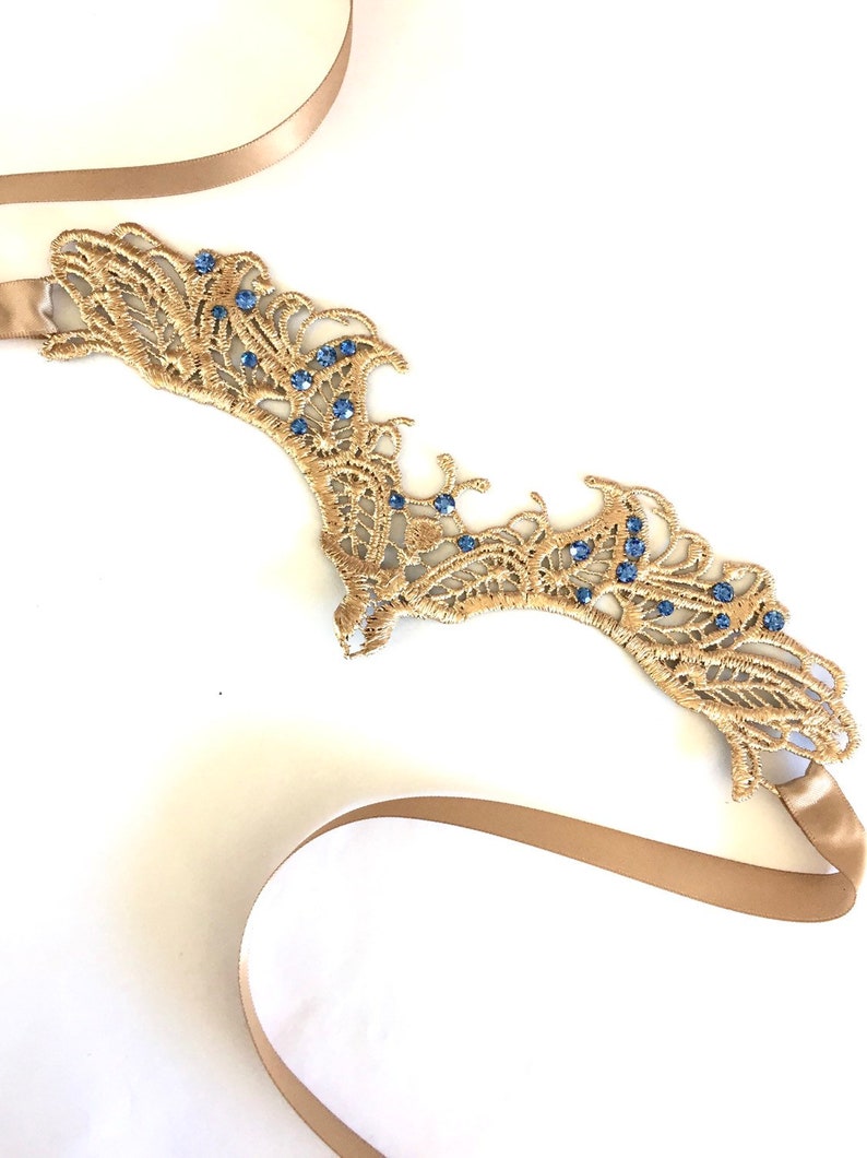 May include: A gold lace headpiece adorned with blue gemstones. The headpiece features an intricate, openwork pattern and is attached to a beige ribbon. This item is likely intended for a special event, such as a wedding or formal occasion.