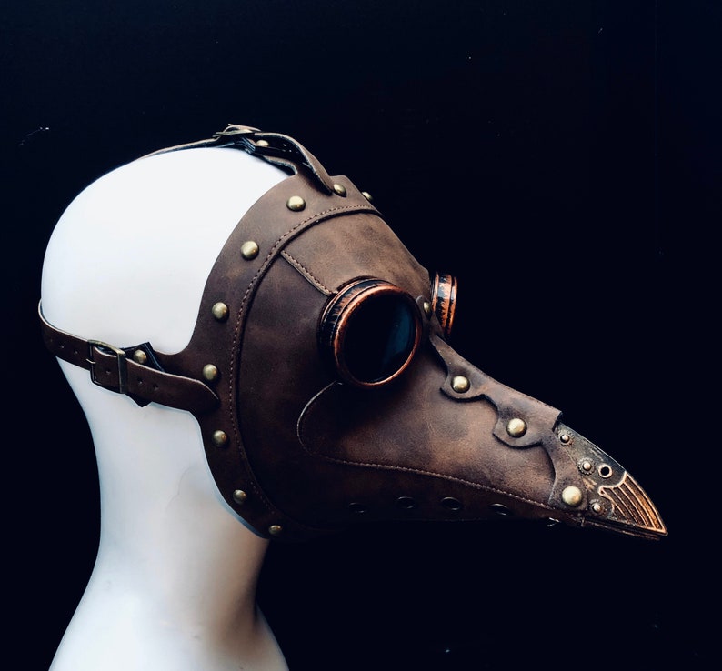 May include: A brown leather plague doctor mask with copper-colored accents and rivets. The mask features a long, beak-like nose, round eye lenses, and adjustable straps. The mask is displayed on a white mannequin head against a black background.