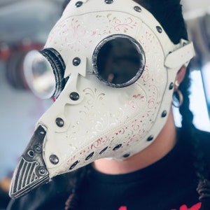 May include: White plague doctor mask with silver-rimmed goggles and a long, beak-like nose. The mask features decorative floral patterns in pink and silver, secured with rivets and straps. The person is wearing a black t-shirt with the word "nightmare" in red.