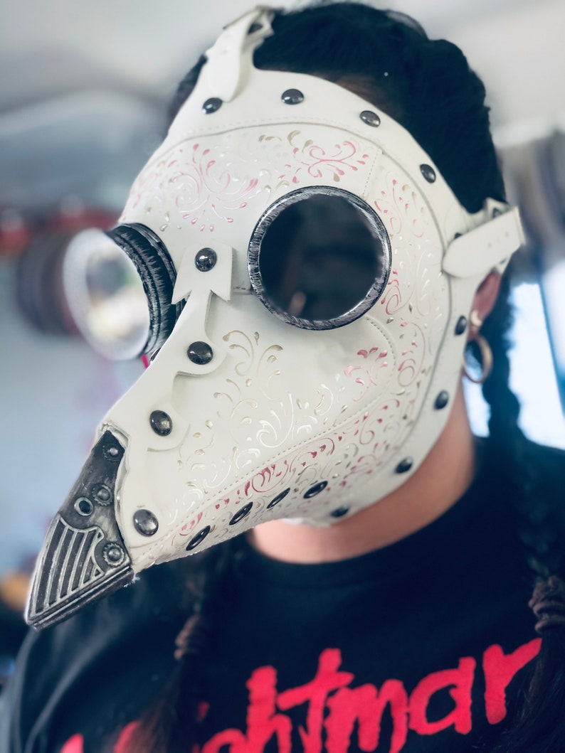 May include: White plague doctor mask with silver-rimmed goggles and a long, beak-like nose. The mask features decorative floral patterns in pink and silver, secured with rivets and straps. The person is wearing a black t-shirt with the word "nightmare" in red.