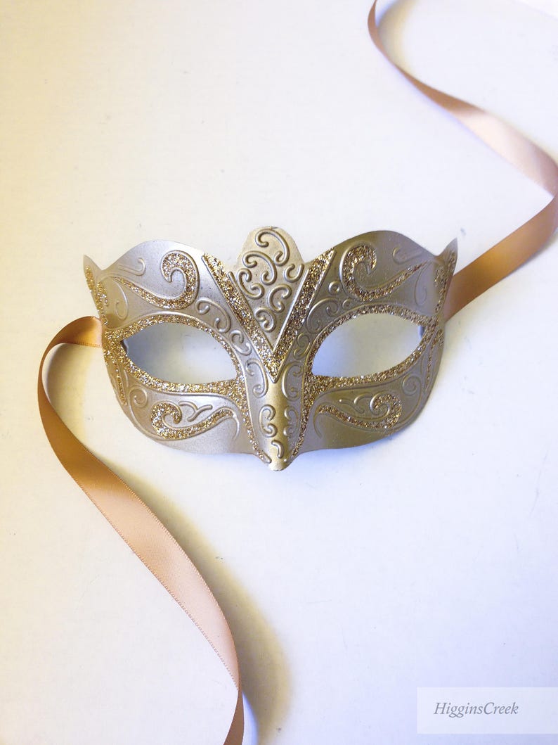 May include: A silver masquerade mask with gold glitter accents and a delicate, ornate design. The mask has a satin ribbon attached for tying.
