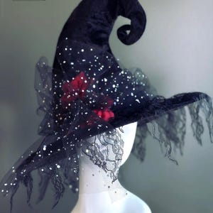 May include: A black witch hat with a velvet finish, adorned with sparkling accents and a delicate lace trim. The hat features a curled tip and a red floral embellishment, perfect for Halloween or costume parties. The Masquerade Store logo is visible in the top left corner.