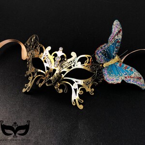 Puede incluir: Una m&aacute;scara de carnaval dorada con detalles intrincados y pedrer&iacute;a, adornada con una mariposa de brillantina azul y roja.