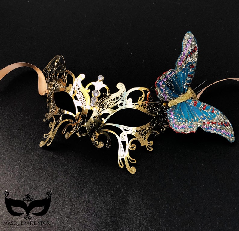 Puede incluir: Una m&aacute;scara de carnaval dorada con detalles intrincados y pedrer&iacute;a, adornada con una mariposa de brillantina azul y roja.