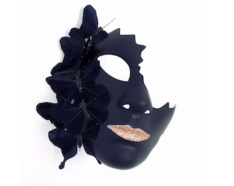 Puede incluir: Una máscara negra de carnaval con un diseño de rostro. La máscara presenta un labio dorado brillante y está adornada con varios adornos de mariposas negras. La máscara tiene un borde dentado en el lado derecho.