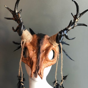 May include: A brown and black skull mask with antlers and black feathers. The mask is made of paper mache and has a rustic look.