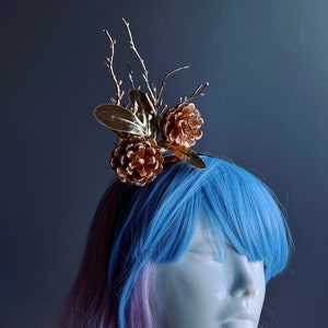 May include: A gold-colored hair accessory featuring pine cones and branches. The accessory is worn by a mannequin with blue and pink hair.