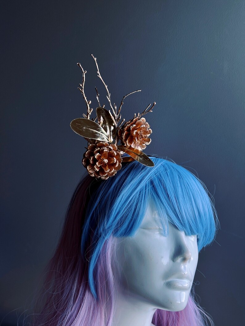 May include: A gold-colored hair accessory featuring pine cones and branches. The accessory is worn by a mannequin with blue and pink hair.