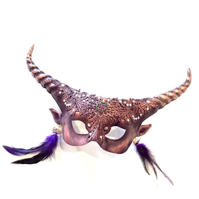 May include: A handcrafted, copper-colored masquerade mask with curved horns and decorative lace detailing. The mask features small ear-like protrusions, embellished with purple feathers and sparkling accents. The horns have a striped pattern, and the mask is designed for a festive occasion.