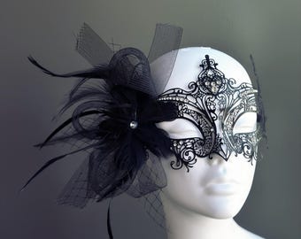 May include: A black masquerade mask with intricate lace detailing and rhinestones. The mask is adorned with a black feather and tulle bow.