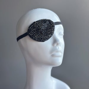 May include: Black, oval eye patch with a beaded design, held in place by a black elastic band. The eye patch is displayed on a white mannequin head. The text "MASQUERADE STORE .COM" is above the mannequin head.