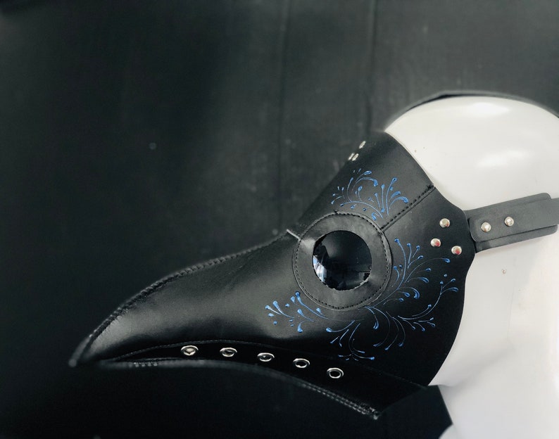 Puede incluir: Una m&aacute;scara de m&eacute;dico de la peste de cuero negro con un dise&ntilde;o floral azul y un agujero redondo para el ojo. La m&aacute;scara tiene una correa y remaches met&aacute;licos.