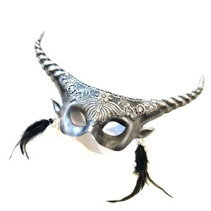 May include: A silver-colored masquerade mask with curved horns and intricate lace detailing. Black feathers dangle from the sides, and the mask has a metallic finish. The mask is designed for a costume or special event.