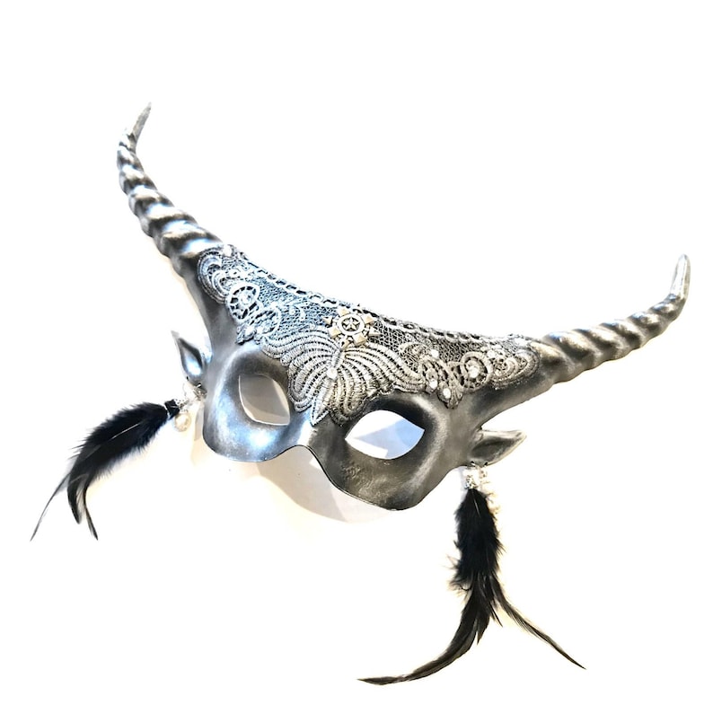 May include: A silver-colored masquerade mask with curved horns and intricate lace detailing. Black feathers dangle from the sides, and the mask has a metallic finish. The mask is designed for a costume or special event.