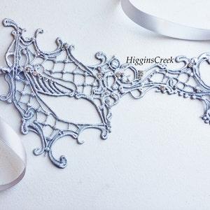 May include: A silver lace masquerade mask with rhinestones. The mask is attached to a silver ribbon.