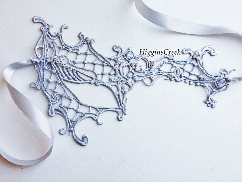 May include: A silver lace masquerade mask with rhinestones. The mask is attached to a silver ribbon.