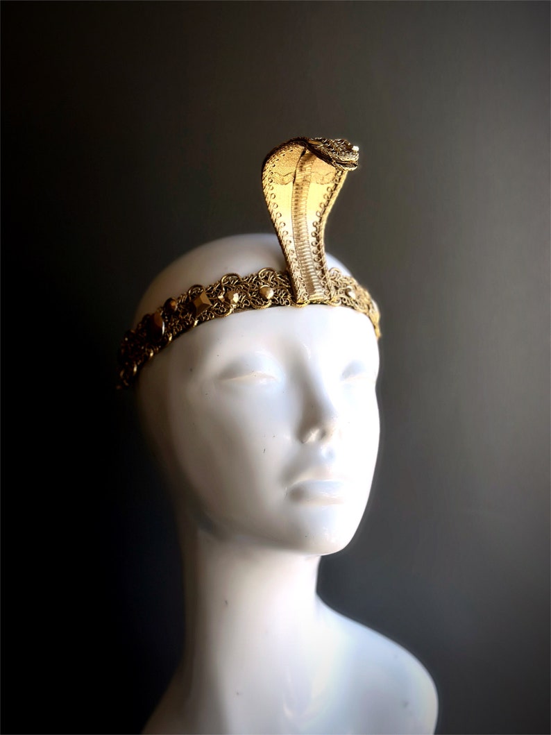 May include: A gold-colored cobra headdress, featuring a detailed cobra head rising from a decorative band. The band is adorned with intricate patterns and small embellishments. The headdress is displayed on a white mannequin head against a dark background.