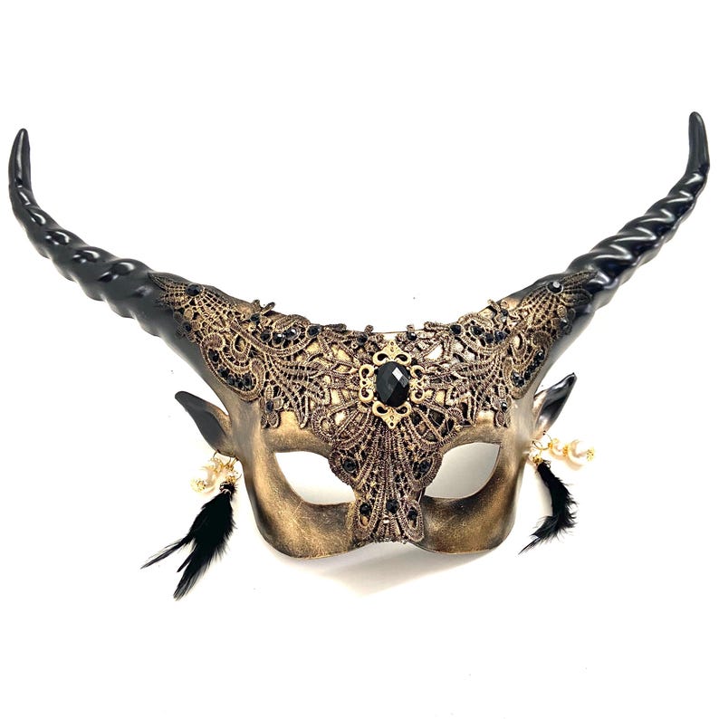 May include: A decorative mask with a gold and black color scheme. The mask features two large, curved black horns, intricate lace detailing, and a central black gem. Black feathers and gold beads dangle from the sides.