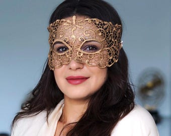 May include: A gold lace mask with a detailed floral pattern. The mask covers the eyes and forehead, featuring delicate designs and a scalloped edge. The mask is worn by a person with dark hair and a white top.