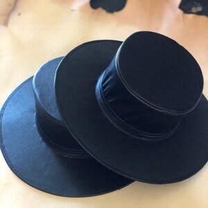May include: Two black plague doctor hats with a wide brim and tall, cylindrical crown. The hats are made of a smooth, black fabric.