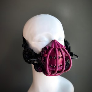 Peut inclure: Un masque de style steampunk rose foncé avec des engrenages et des tubes noirs. Le masque a une forme arrondie et est conçu pour couvrir le nez et la bouche.