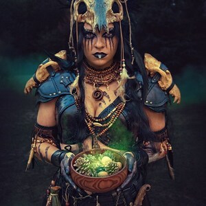 May include: A woman wearing a horned headdress and leather armor holds a wooden bowl filled with pine cones and a glowing green liquid. She has dark hair, dark eyes, and a dark lip ring.