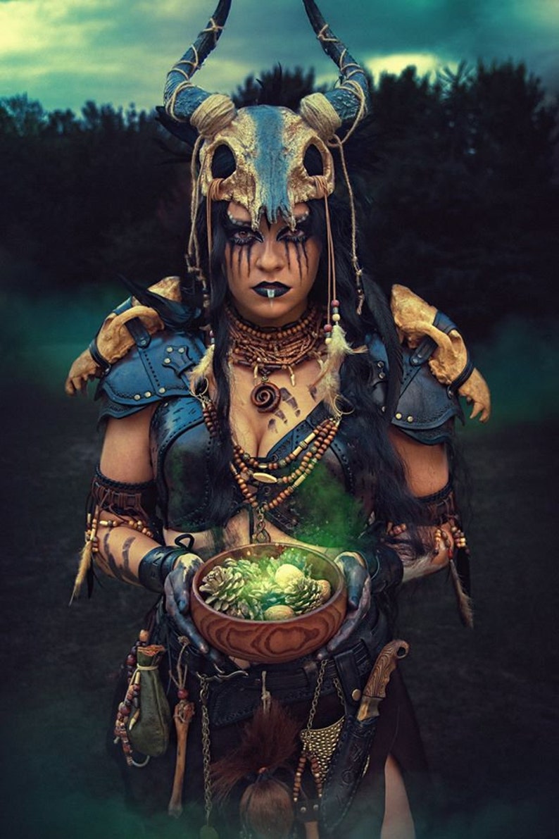May include: A woman wearing a horned headdress and leather armor holds a wooden bowl filled with pine cones and a glowing green liquid. She has dark hair, dark eyes, and a dark lip ring.
