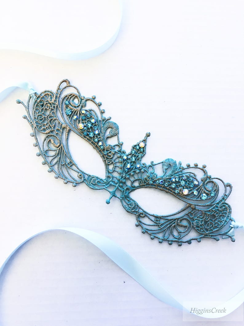 May include: A teal blue masquerade mask with intricate lace detailing and sparkling rhinestones. The mask is attached to a light blue satin ribbon.