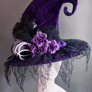 Puede incluir: Sombrero de bruja de terciopelo morado con ala ancha, ribete de encaje negro y rosas y plumas moradas. El sombrero tiene una copa puntiaguda y un velo de encaje.