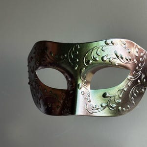 May include: A decorative eye mask with an ornate floral design. The mask is a metallic color, with shades of green and bronze. The mask has two eye holes and is designed to be worn at a masquerade ball or costume party.