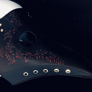 Puede incluir: Una m&aacute;scara de m&eacute;dico de la peste de cuero negro con un dise&ntilde;o floral rojo y una pieza ocular transparente. La m&aacute;scara tiene un pico largo y se asegura con correas.